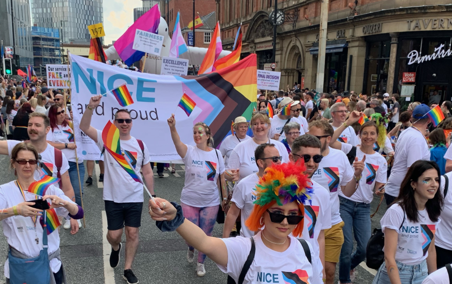 Members of NICE and proud walking during the Manchester pride parade, waving flags and carrying a NICE banner.